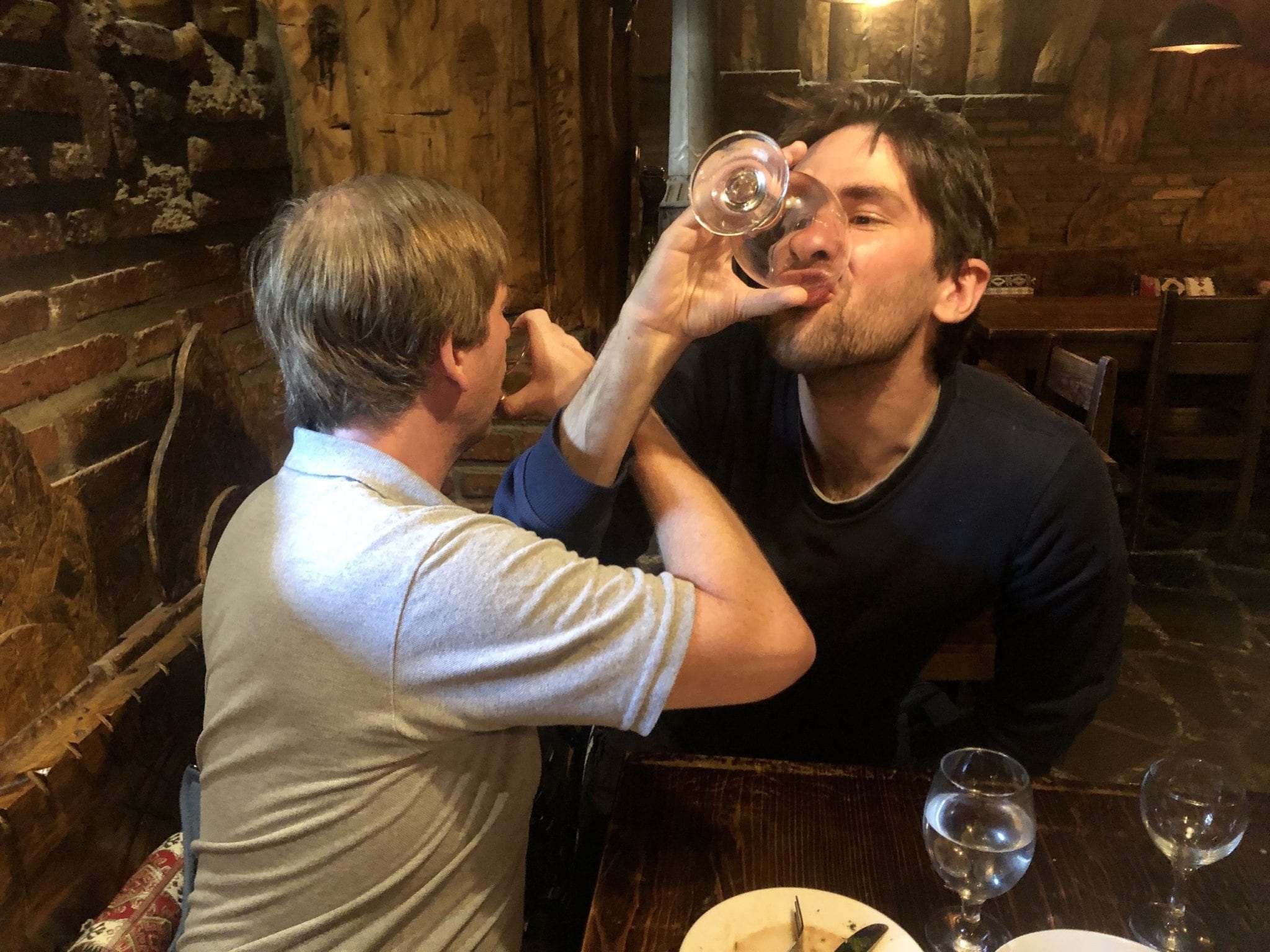 Tom and the Russian owner of the restaurant drink very full glasses of wine with their drinking arms entwined.