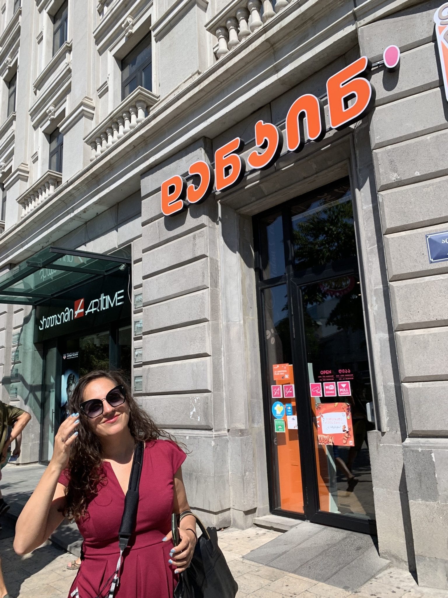 Kate smiles while standing in front of what looks like a Dunkin Donuts, with the name written in Georgian script.