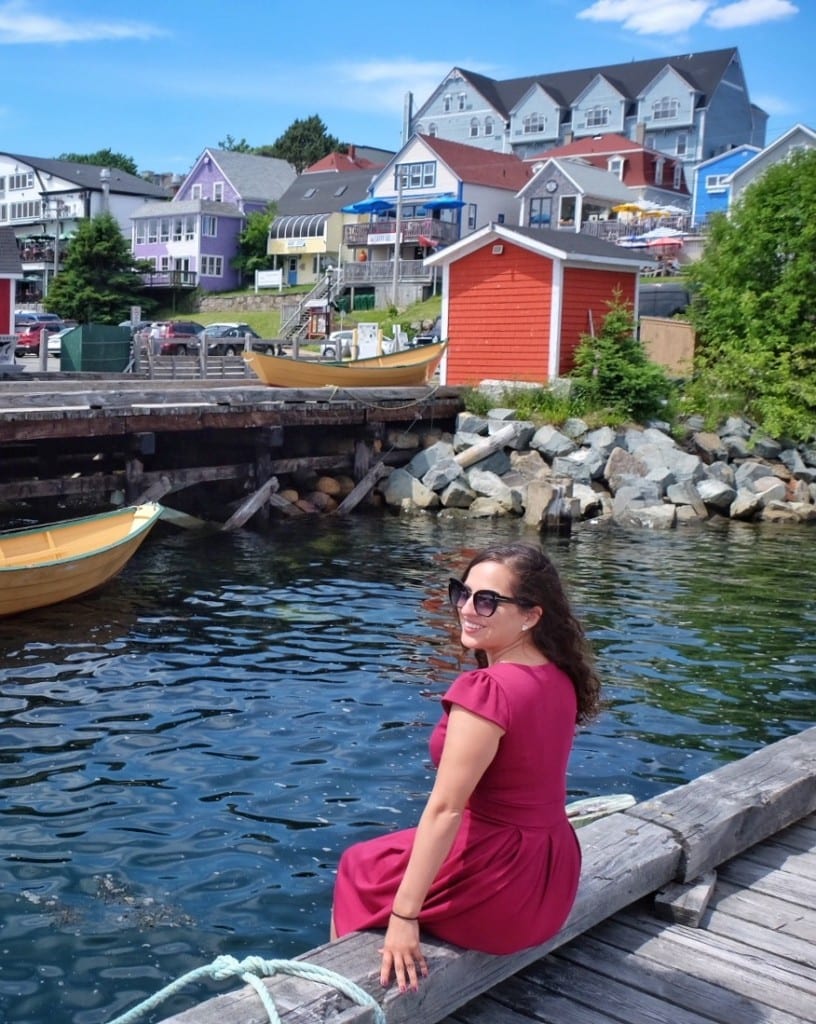 Kate wears a red dress and sits on the end of a wooden pier with old-fashioned wooden homes in the distance behind her.