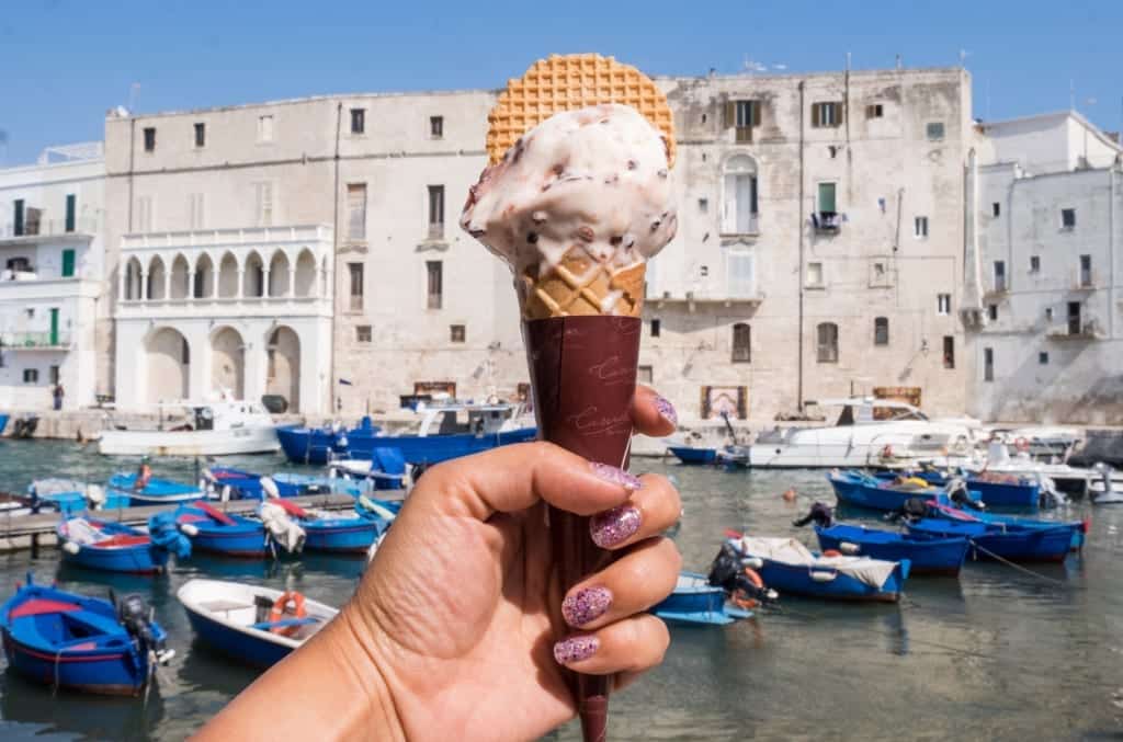 A hand holding up an ice cream with white buildings and blue boats in the background.
