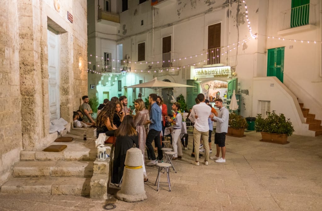 People hanging out in the street while enjoying drinks, green-shuttered buildings and stringed lights in the background.