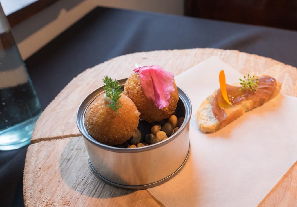 Tiny fried crab balls in a silver cup, next to a piece of crostini with cured lamb.