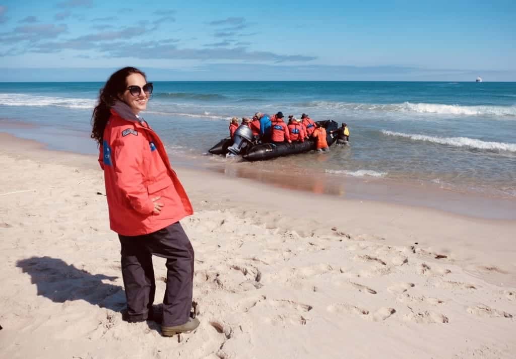Kate wearing a red coat and standing on the beach in Sable Island, turning her head back to face the camera with a grin, as a zodiac filled with passengers leaves the island.