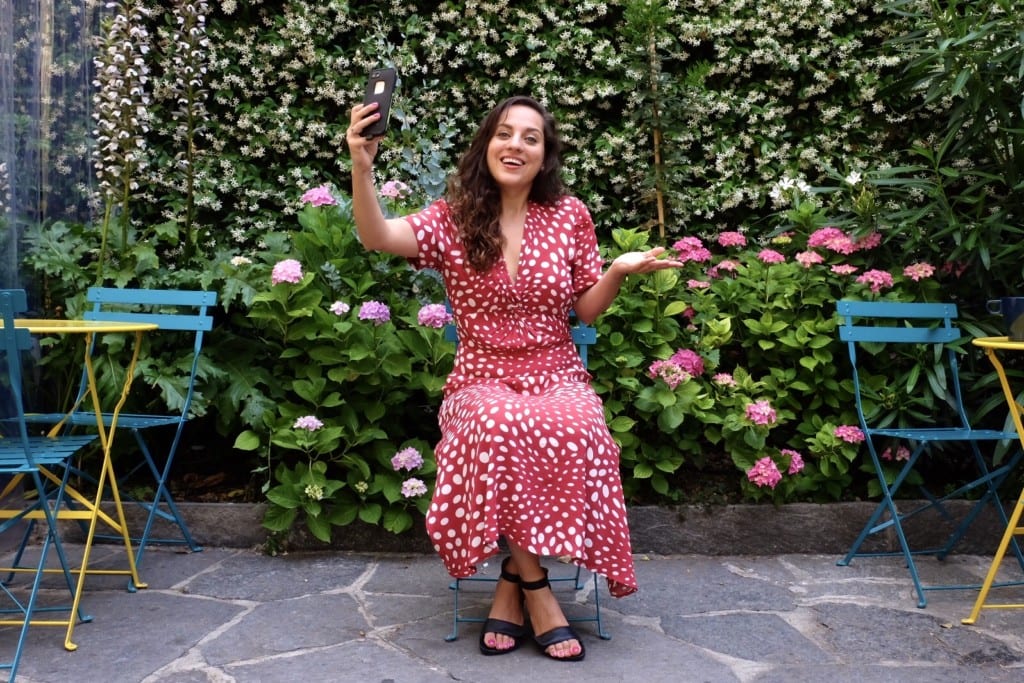 Kate wears a red and white polka-dot dress and attempts to take a selfie in a garden.