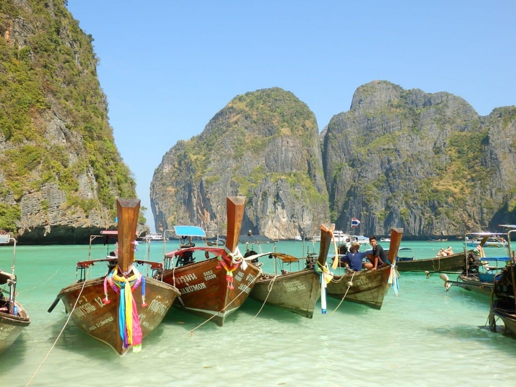 Longtail boats with brightly colored scarves in shallow turquoise water with cliffs in the background.