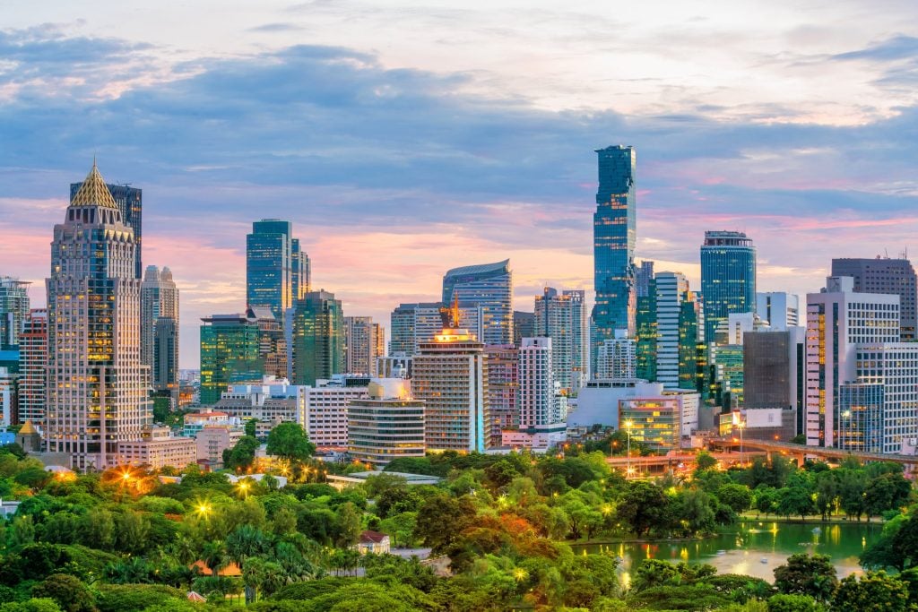 Bangkok's Lumphini Park with rows of skyscrapers behind it, lit up at sunset.