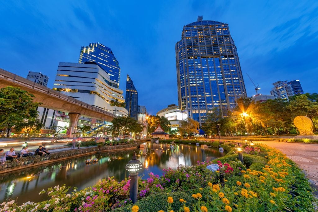 A brightly lit park with a pond at blue hour in Bangkok, busy streets and skyscrapers in the background.