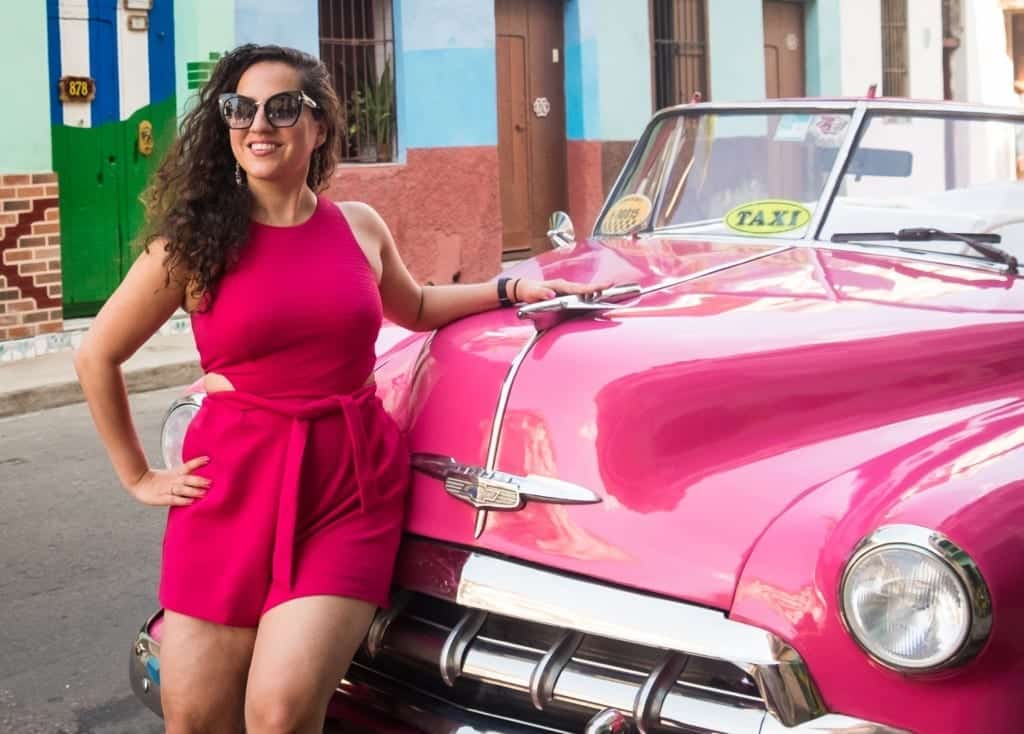 Kate wears a bright pink romper and stands in front of a bright pink classic car in Havana.