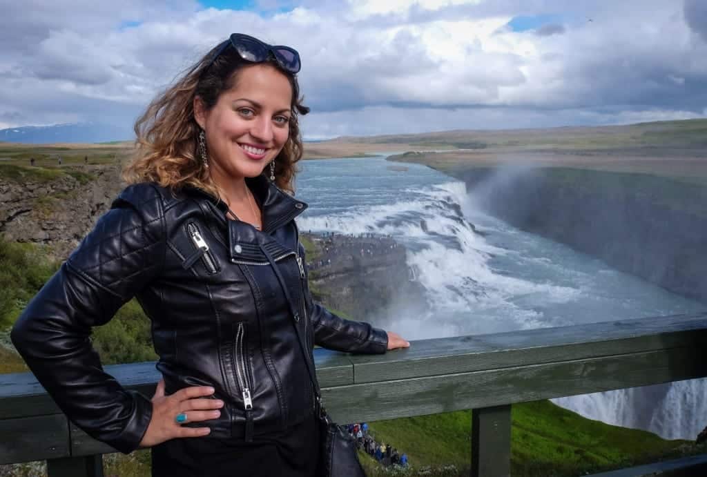 Kate stands in front of Gulfoss waterfall in Iceland, wearing a leather jacket and grinning.