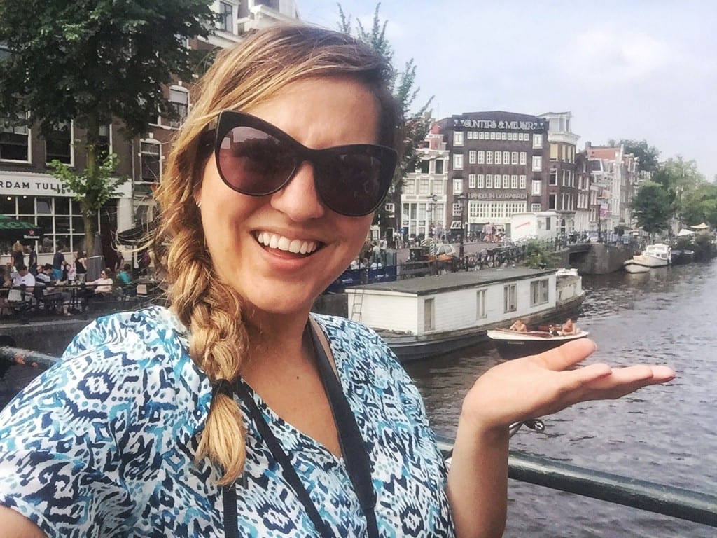 Kate with a blonde braid and sunglasses, holding her hand up as if to say, "What?" with the buildings of Amsterdam and canals in the background.