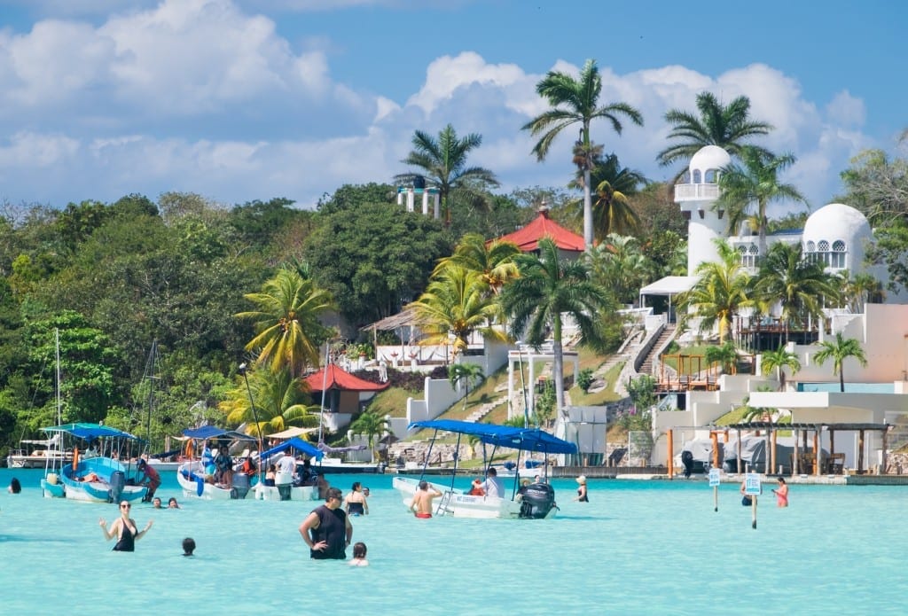 People playing in the pale blue water in front of a castle-like complex on shore.