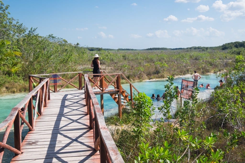 A woman in a black dress and a straw hat on a wooden platform leading to a turquoise river.