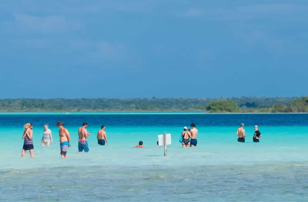 People standing in the shallow water of Bacalar. You can see the colors change from pale aquamarine to bright turquoise, bright blue and dark inky navy.