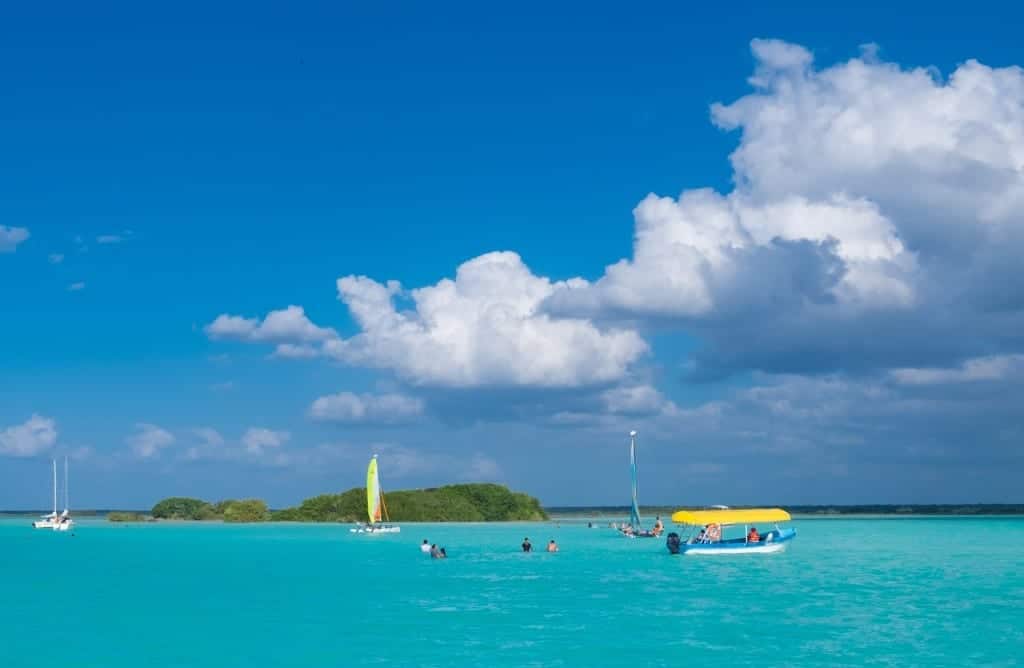 A neon blue section of a lake, people swimming and sailing around a small island.