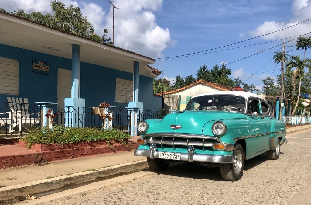 A bright turquoise classic car parked on a street in front of a blue home in Viñales, Cuba.