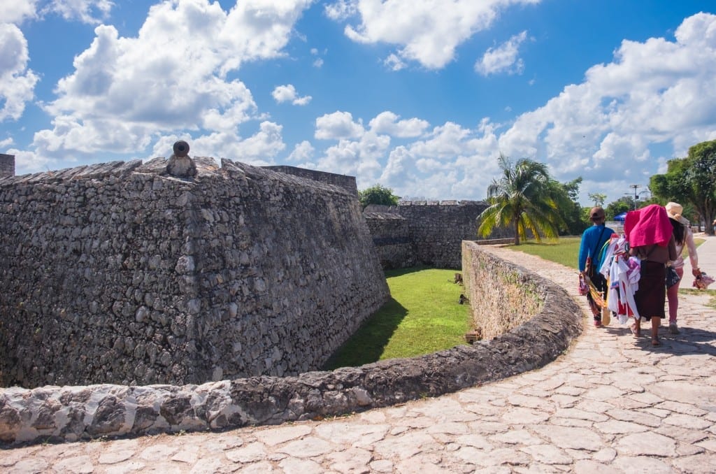 A few street vendors walking past a stone fort.