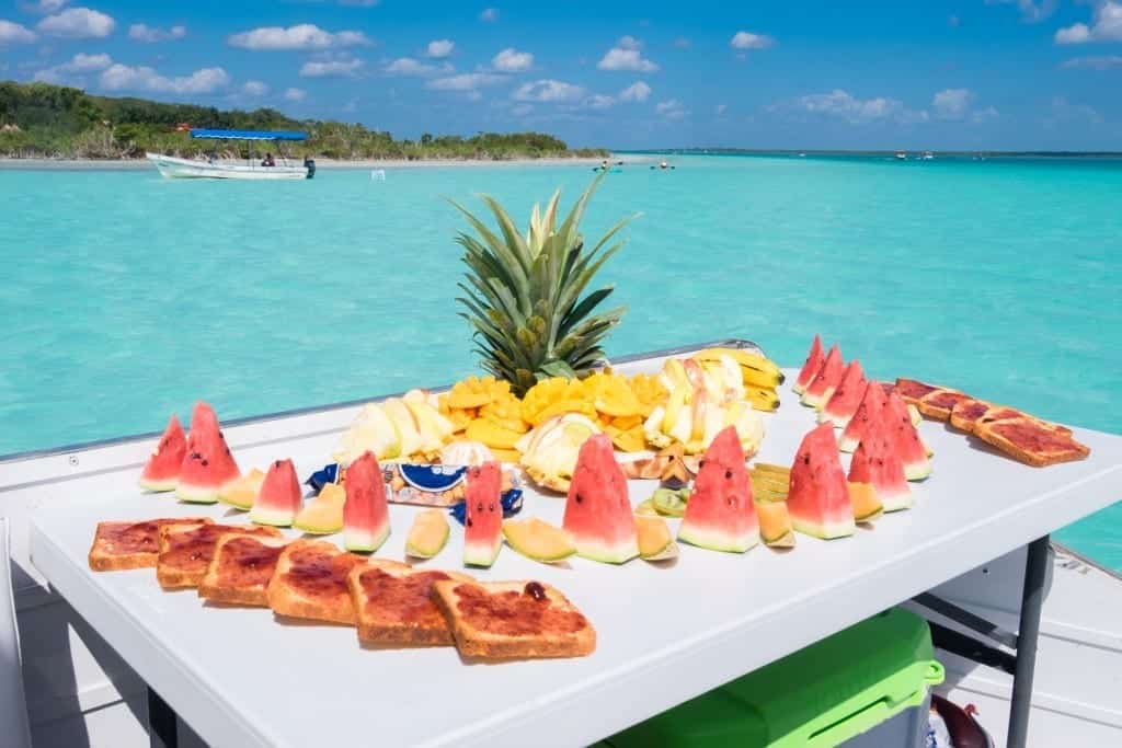A big decorated fruit arrangement of watermelon, pineapple, mango, and cantaloupe (and also some toast with jelly on it for some reason) on a boat on the lake.
