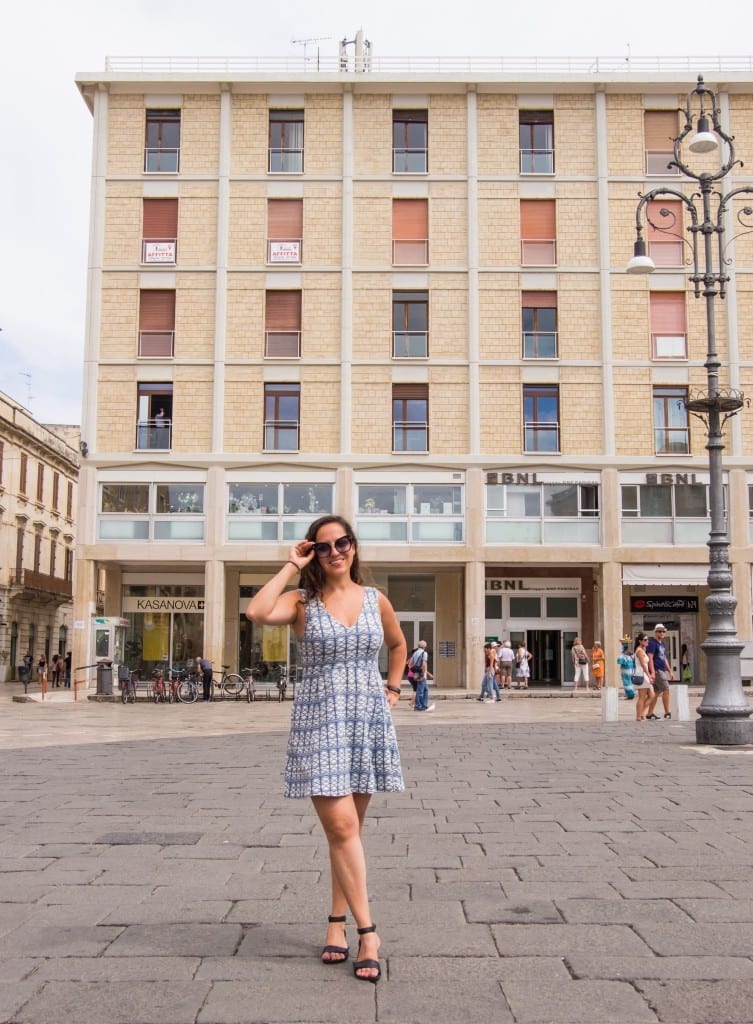 Kate wears a green and white patterned dress and stands in front of a boxy orange building in Lecce.