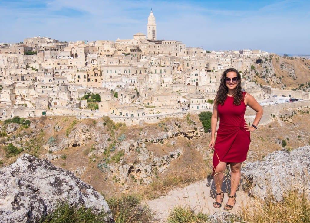 Kate porte une robe rouge à l'ourlet asymétrique et pose devant la ville de Matera : des tours et des maisons en pierre construites sur une rangée de sassi (grottes).
