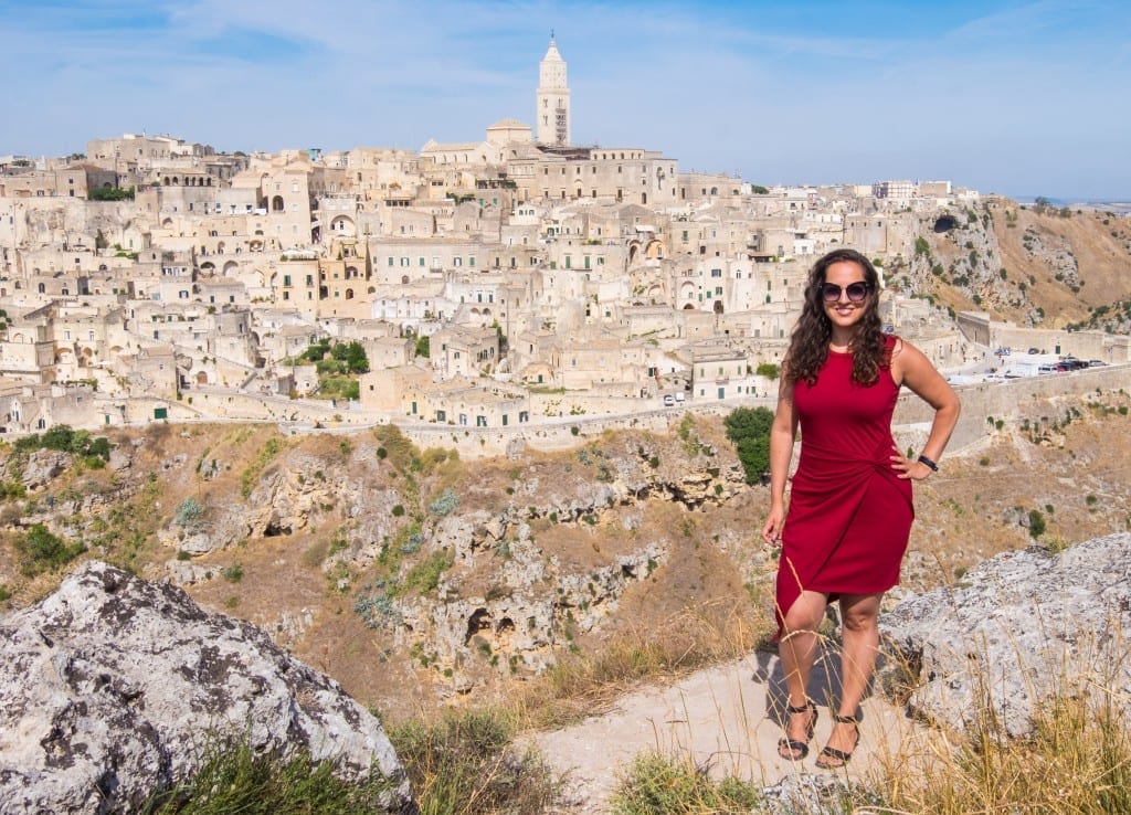 Kate wears a red dress with an asymmetrical hemline and poses in front of the city of Matera: stone towers and homes built on top of a row of sassi (caves).