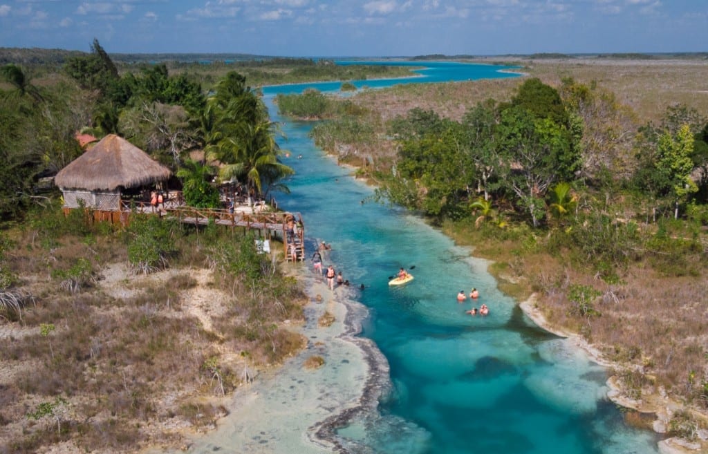 An aerial view of people swimming and kayaking in a narrow, neon turquoise river.