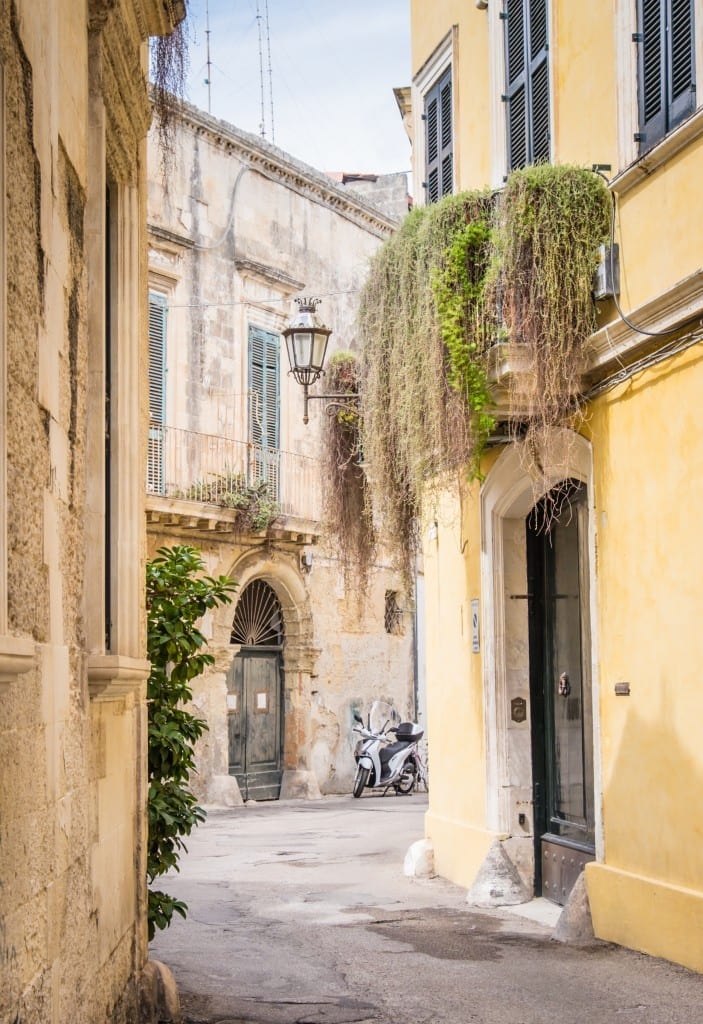 A narrow alley in Italy, with yellow-painted buildings with green doors and shutters, a lush green plant spilling from a balcony, a motorcycle parked outside.