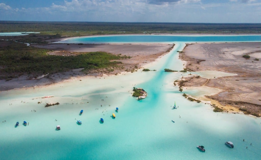 An aerial view of a narrow channel with sand and pale turquoise water leading to a wider river. Lots of sailboats and people in the water.