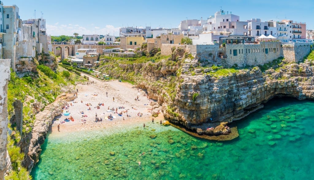 The city of Polignano a Mare, with a sandy white beach leading to clear teal water, with two cliffs on each side, one cliff topped with whitewashed buildings.
