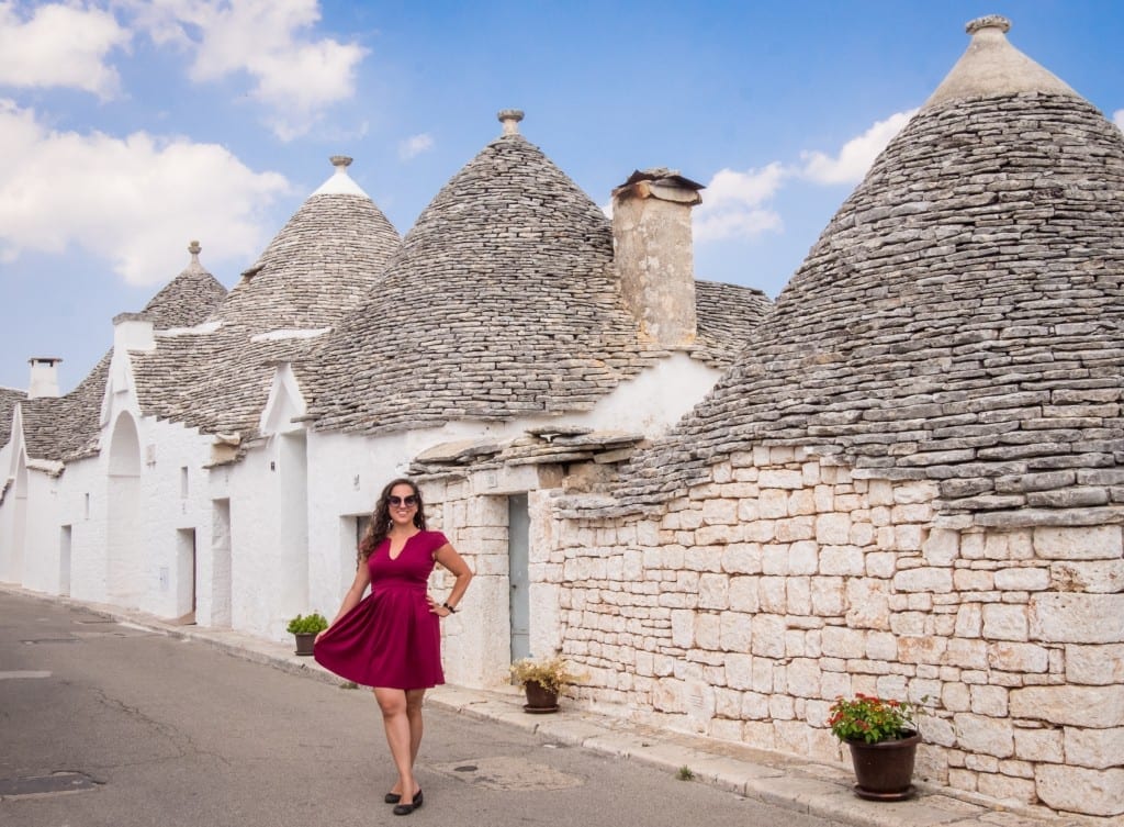 Kate wears a red dress and poses, holding it open, in front of the white conical trulli homes of Puglia.