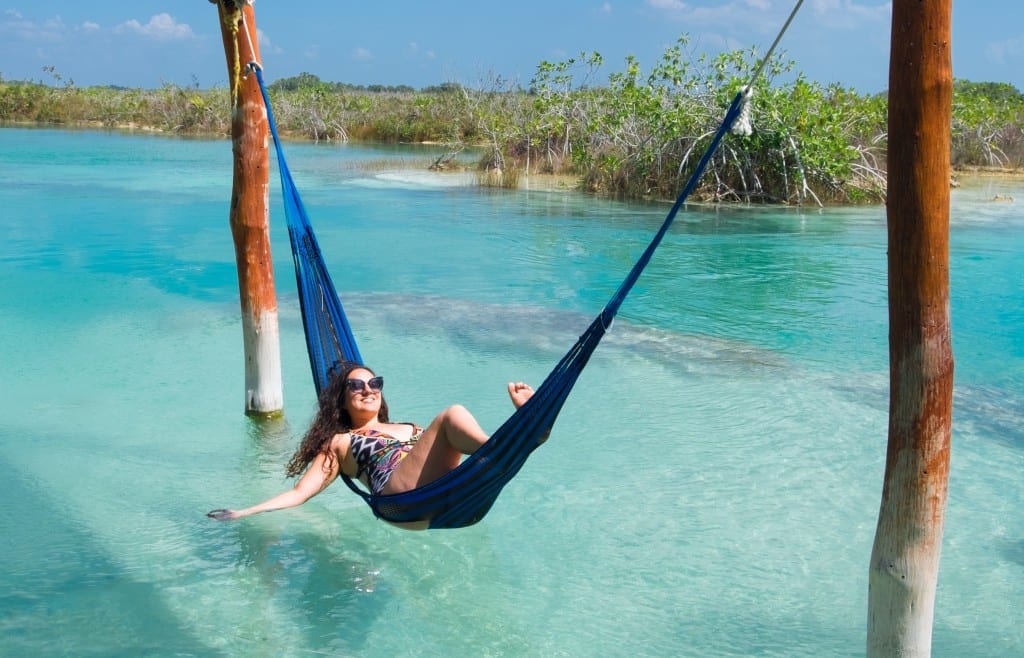 Kate relaxes in a hammock on top of bright clear turquoise water in Bacalar, Mexico, running her hand through the water.