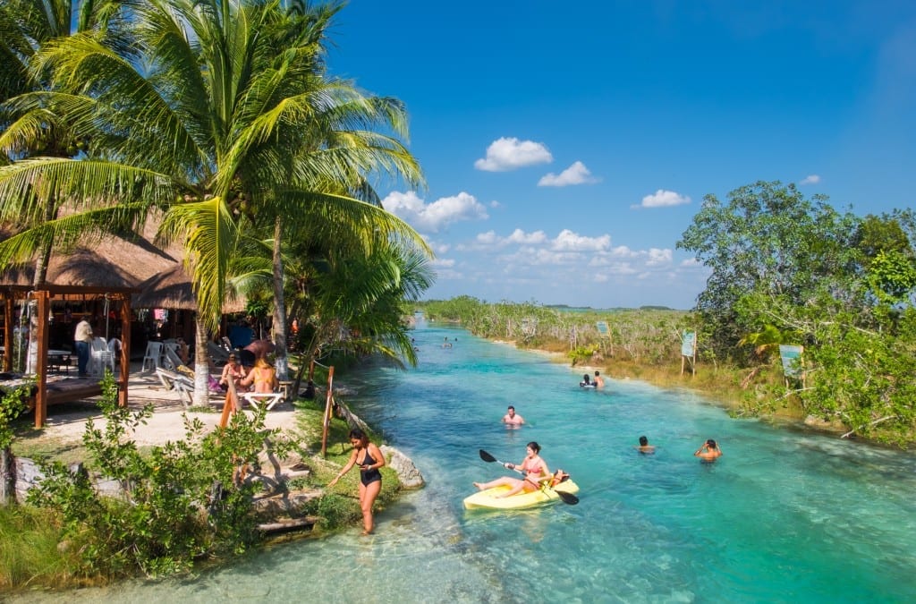 A bright turquoise lazy river next to a restaurant, people swimming and kayaking.