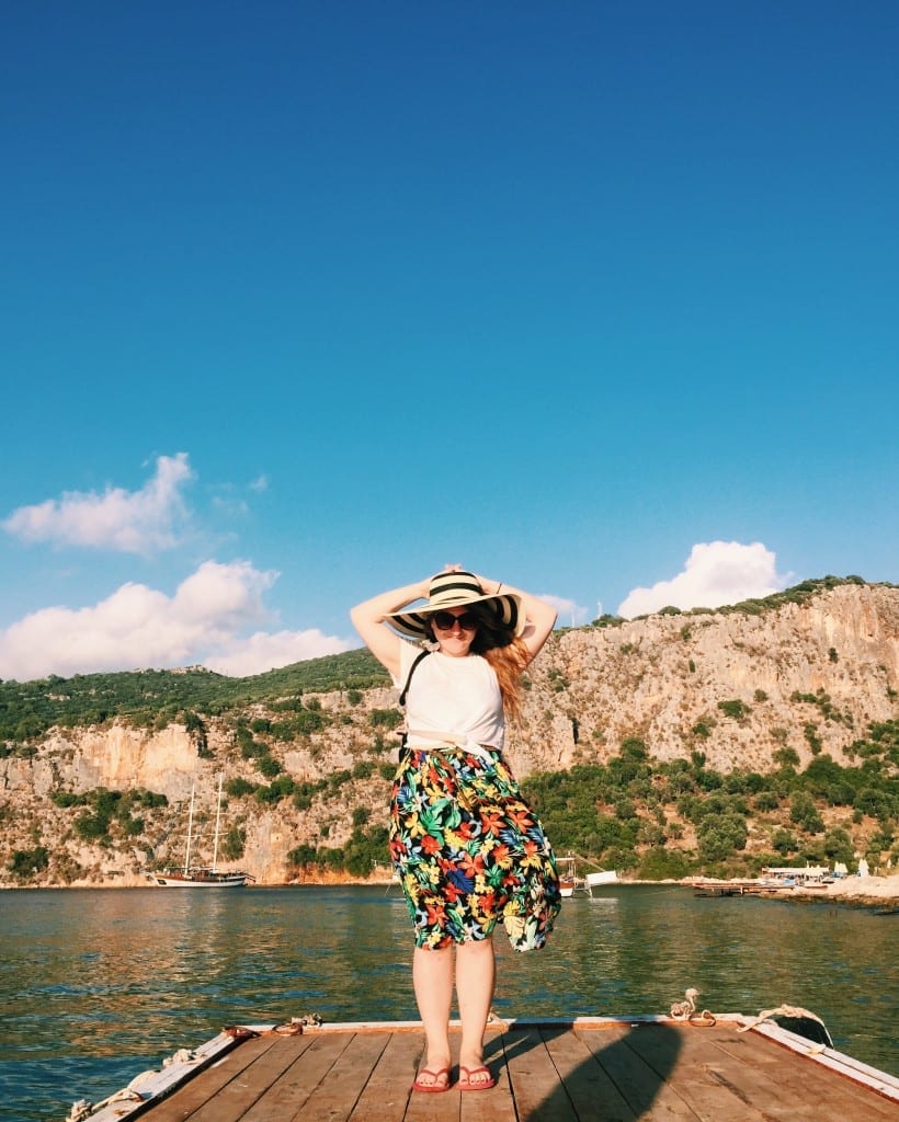 Katie in a patterned skirt, white top, and floppy hat smiling in front of the dark green Mediterranean near Kas.
