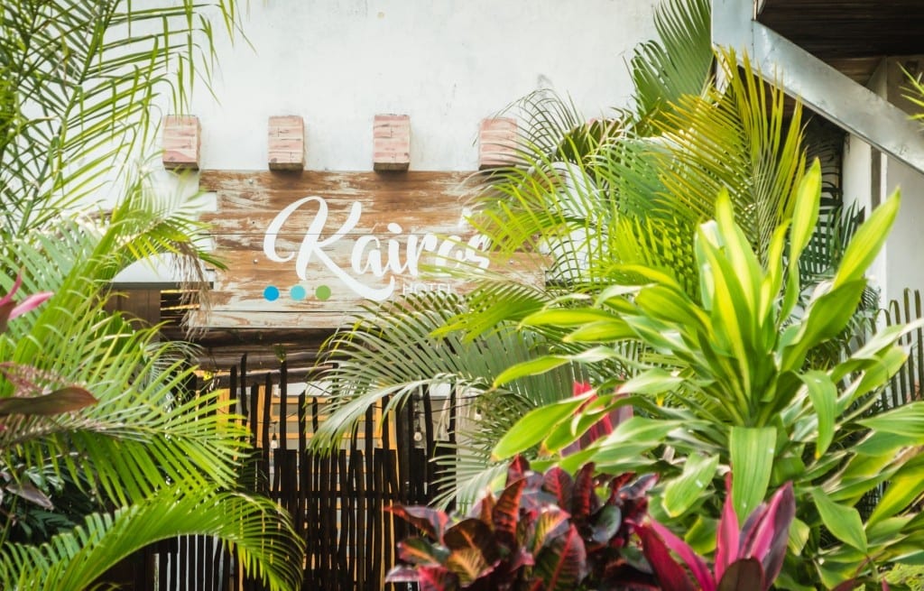 A sign reading Kairos Hotel surrounded by palm trees.