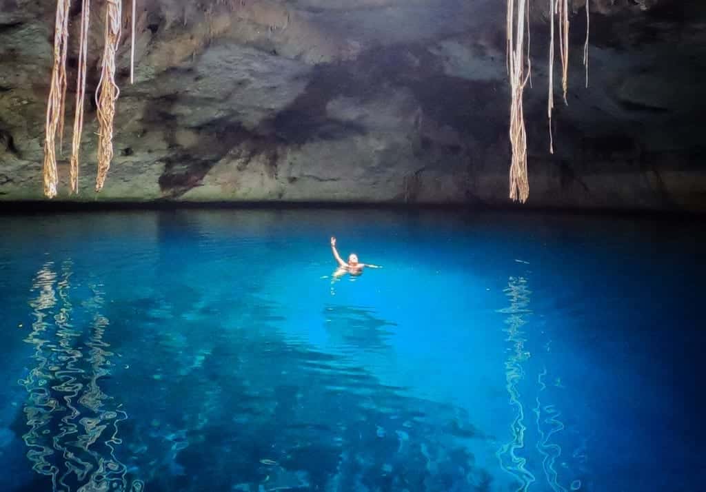 Kate swimming in the bright electric blue Cenote of Noh Mozon, raising her arm in the air.