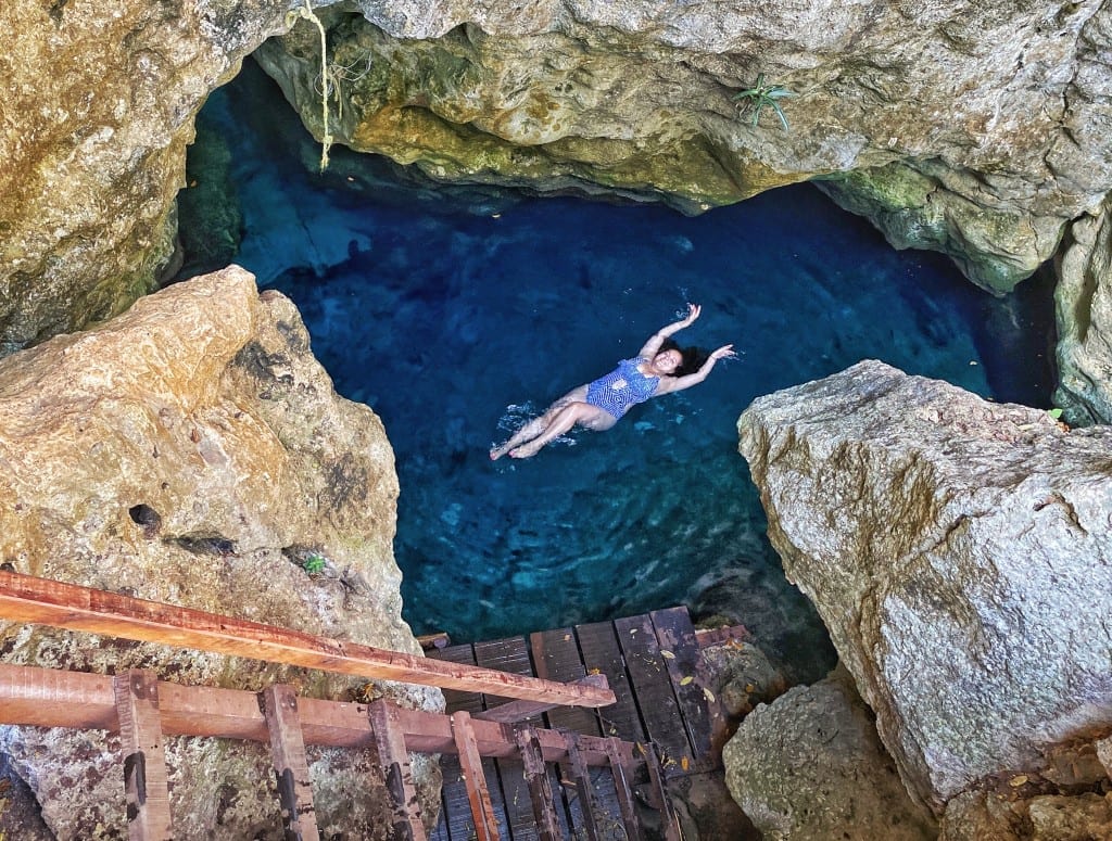 Kate floats in a tiny bright blue cenote, surrounded by rocks and a wooden staircase.