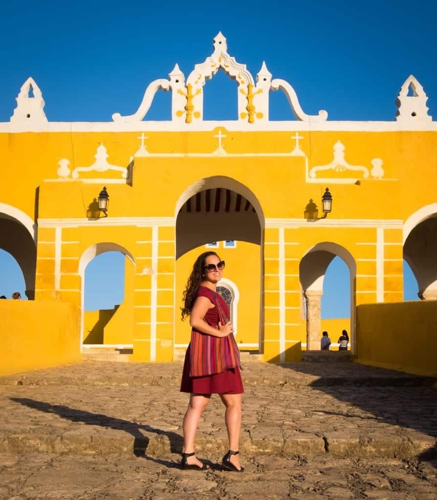 Kate wearing a red dress, standing in front of a bright yellow and white gate of Izamal in front of a bright blue sky. She holds a multicolored striped purse around her shoulder.