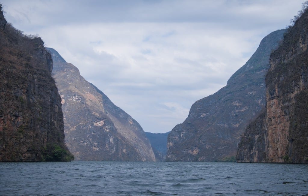 A still blue river surrounded by encroaching orange and green canyon walls.