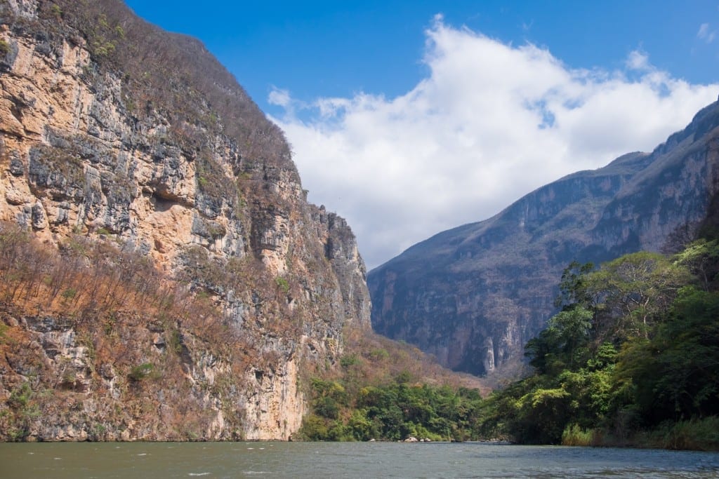 The walls of the canyon rise up against a blue and white sky, the river bright green.