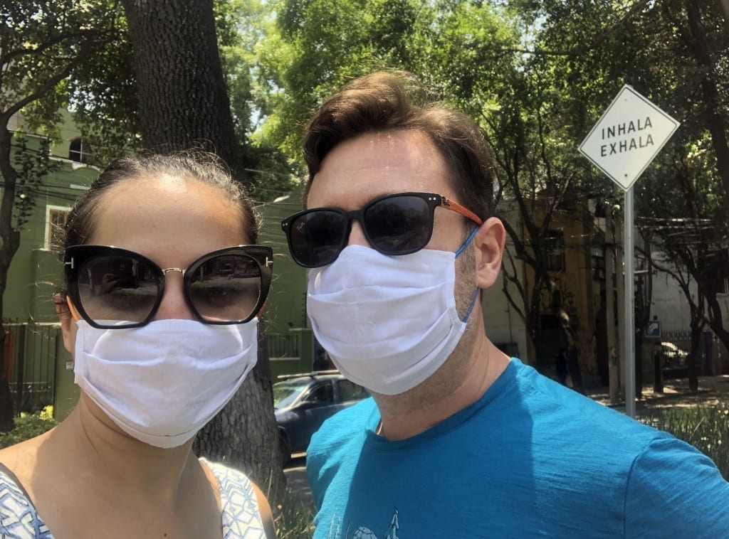 Kate and Charlie wearing face masks and sunglasses in front of a sign that reads "Inhala Exhala" in Mexico City.