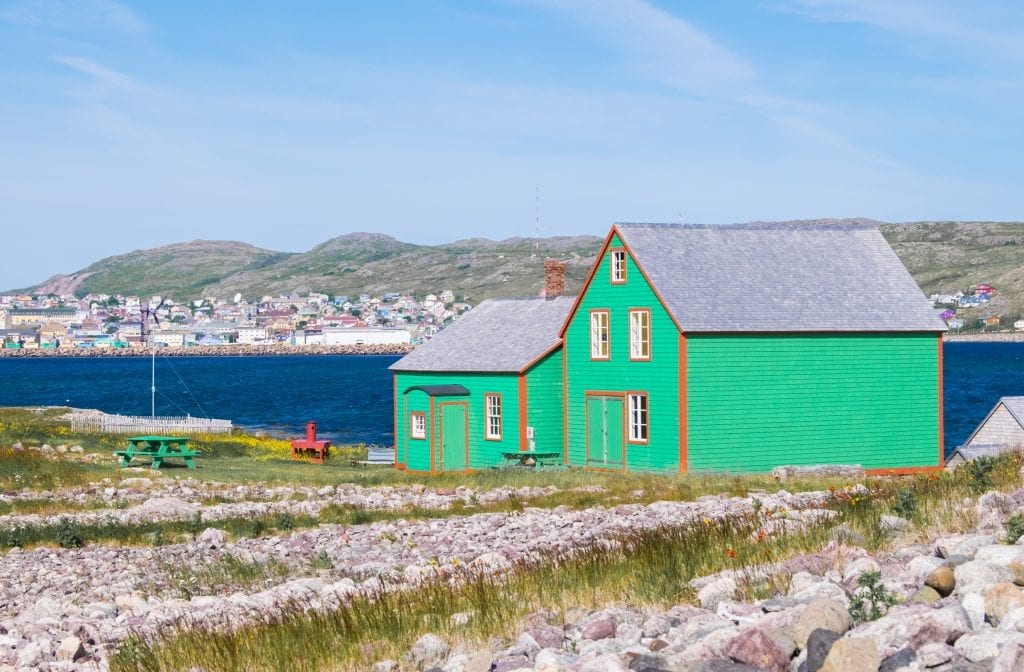 A bright green cottage with orange trim on the edge of the ocean, St. Pierre tow in the background.