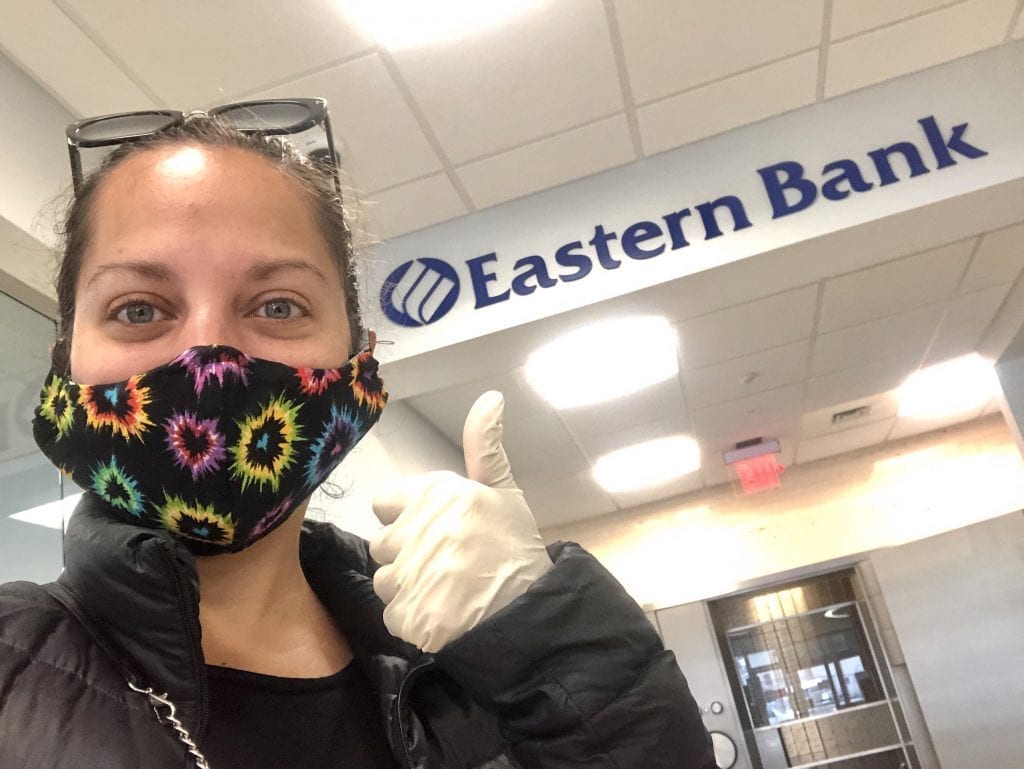 Kate poses with a mask on by an "Eastern Bank" sign, giving the thumbs up with a rubber gloved hand.