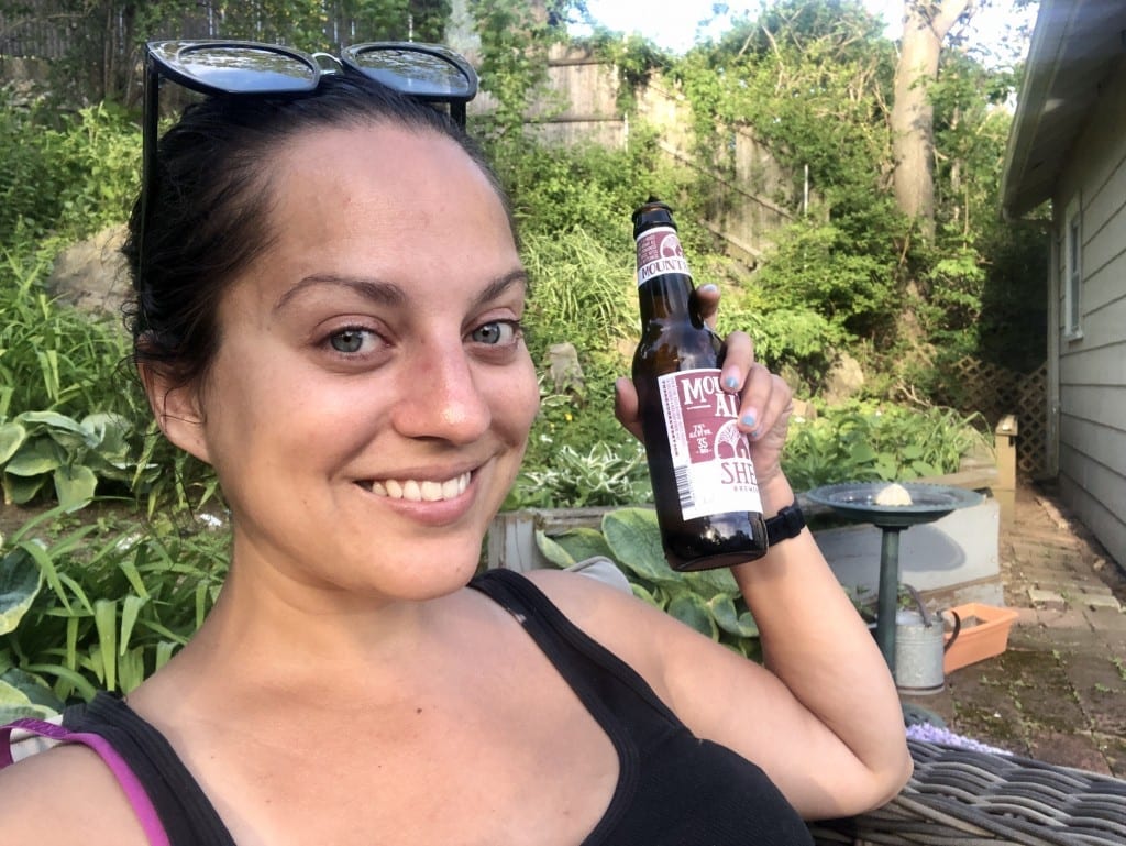 Kate sits outside in a garden area, holding up a bottle of beer and smiling.
