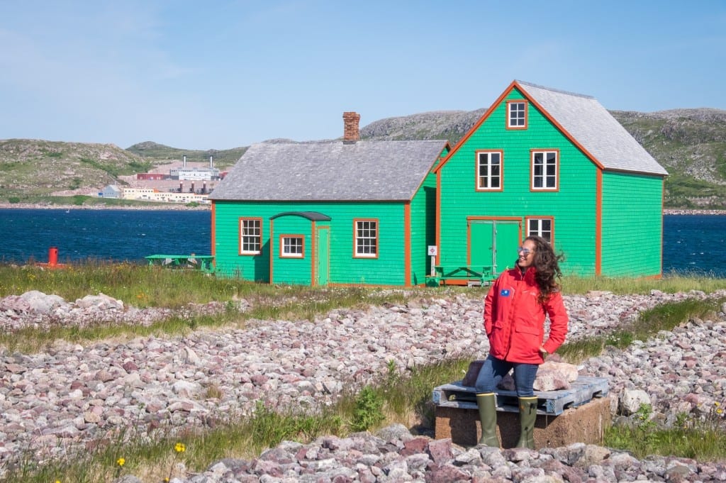 Kate wears a bright red jacket and stands in front of a green wooden cottage on the rocky shore of St. Pierre island.