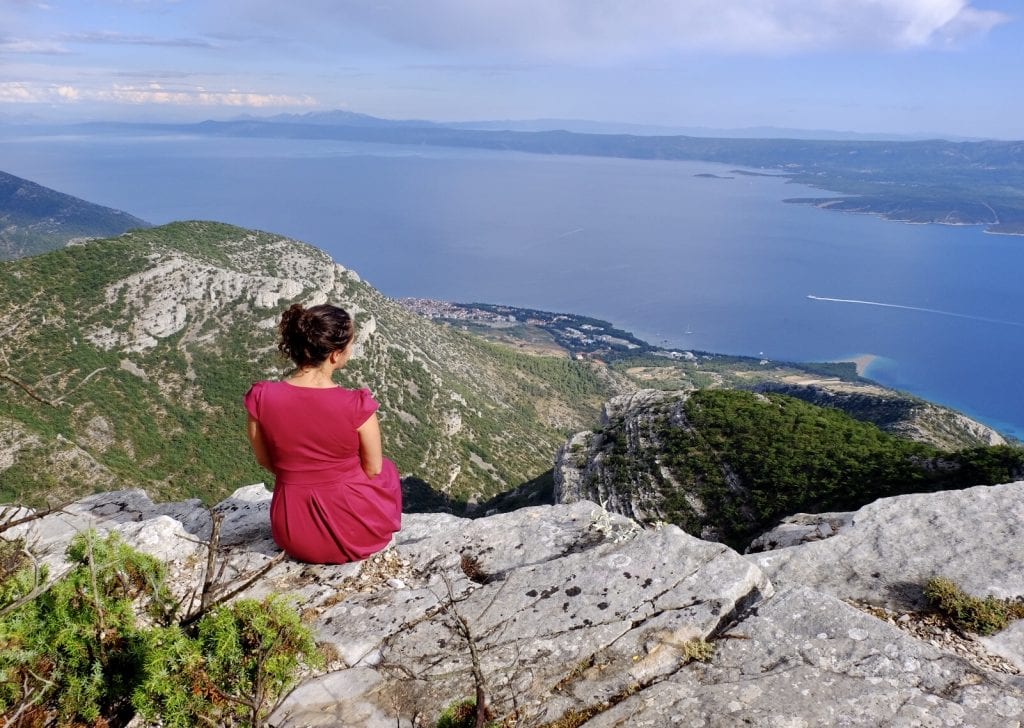 Kate wears a red dress and sits on the edge of a mountain overlooking the view to the sea in Brač, Croatia.