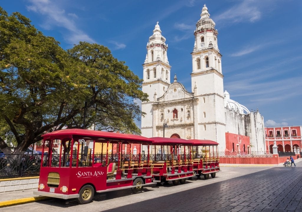 A red streetcar-style bus parked in front of a white colonial-era church in front of a park, set against a bright blue sky.