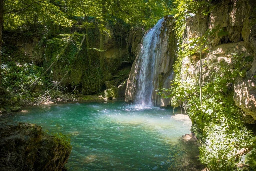 A waterfall surrounded by lush greenery emptying into a blue-green pool in the woods.