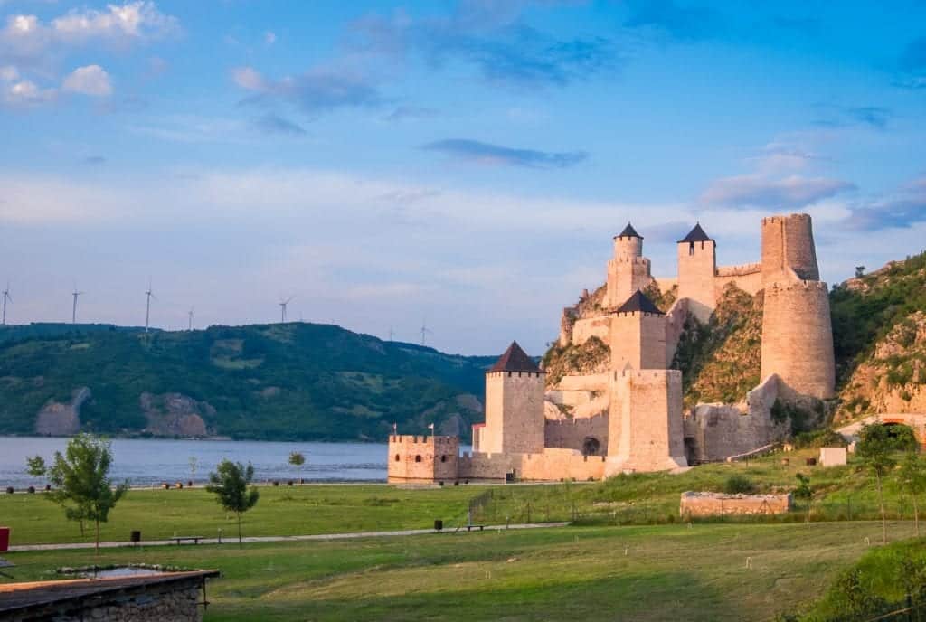 Golubac Fortress, which looks like a Harry Potter-like place with towers sticking out of a hillside in front of the Danube. The fortress is lit up orange from the sunset underneath a bright blue sky.