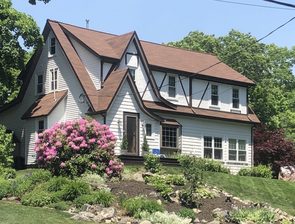 A white house with brown trim and lots of pointy eaves, a purple rhododendron bush in front.