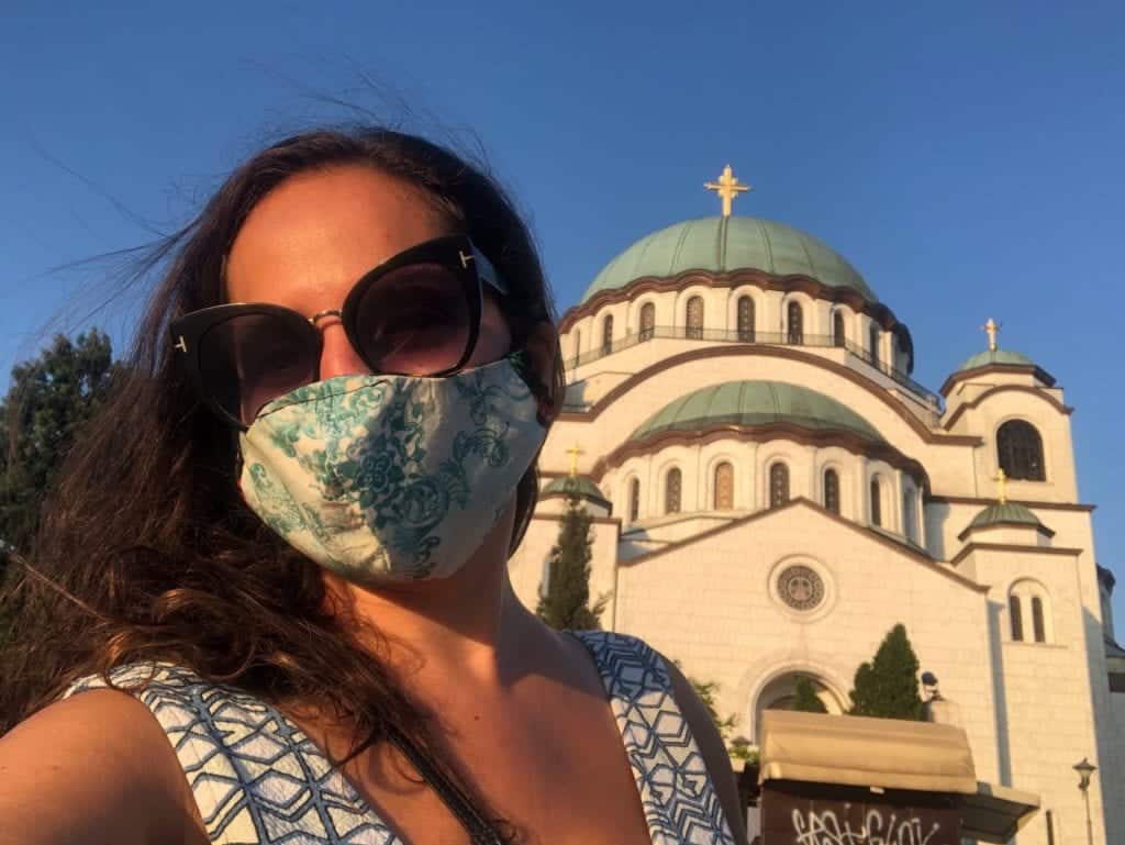 Kate wears a green and white dress and a green and white face mask with sunglasses, in front of the green and white church in Belgrade underneath a bright blue sky.