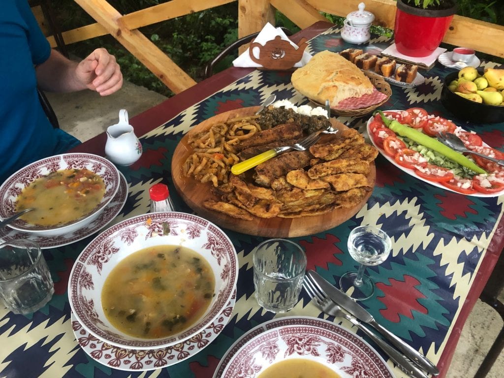 Tons of food on a patterned tablecloth, including big bowls of fish soup, fried fish and zucchini, spinach, kajmak, boiled beans, a platter of tomatoes topped with onions and cucumbers with a big green pepper on top, a loaf of bread, a bowl of small pears, four slices of plum-stuffed cake.